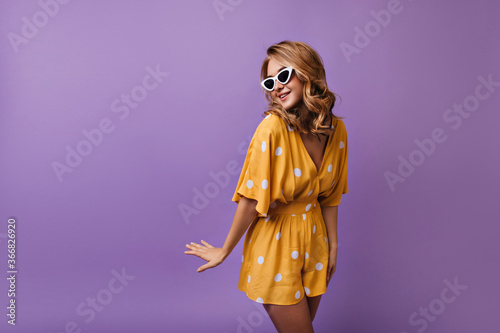Stunning european lady with tanned skin posing with pleasure on purple background. Attractive blonde girl in summer outfit dancing in studio.
