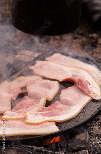 Middle bacon rashers cook on an iron plate, over an open campfire - smoke and steam fill the chilly morning air - with a black billy hanging above. 