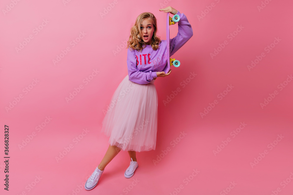 Full-length shot of surprised girl in lush skirt holding longboard. Indoor photo of amazing young woman with skateboard.