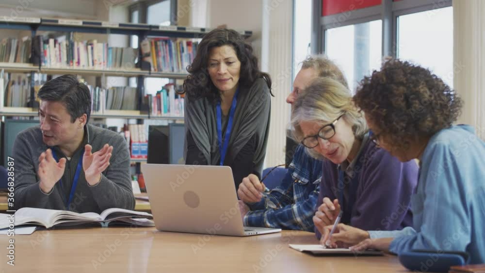 Female teacher helping group of mature multi-cultural students studying and working on laptop in library learning IT skills in evening class - shot in slow motion