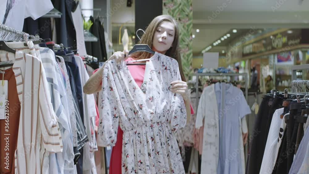 Young pretty woman is trying floral printed dress while standing between clothes racks in the shop. Attractive girl is looking at herself to the camera like in mirror, turning and smiling. Young pretty woman is trying floral printed dress while standing between clothes racks in the shop. Attractive girl is looking at herself to the camera like in mirror, turning and smiling.