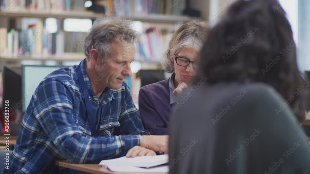 Female teacher helping mature male student studying and using mobile phone in library learning IT skills in evening class - shot in slow motion Female teacher helping mature male student studying and using mobile phone in library learning IT skills in evening class - shot in slow motion