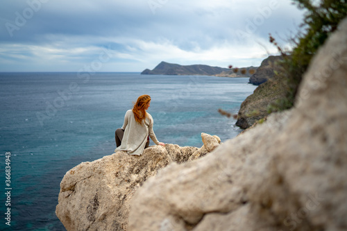 Rear view of woman sitting on rock by sea against cloudy sky