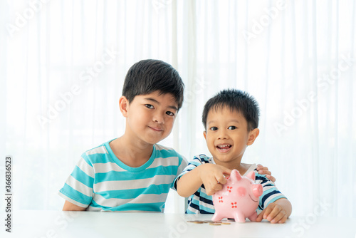 Two Asian cute brother and son insert coin  in piggy bank together on the table in living room at home. Concept of bonding of sibling, friendship and saving, investment money concept