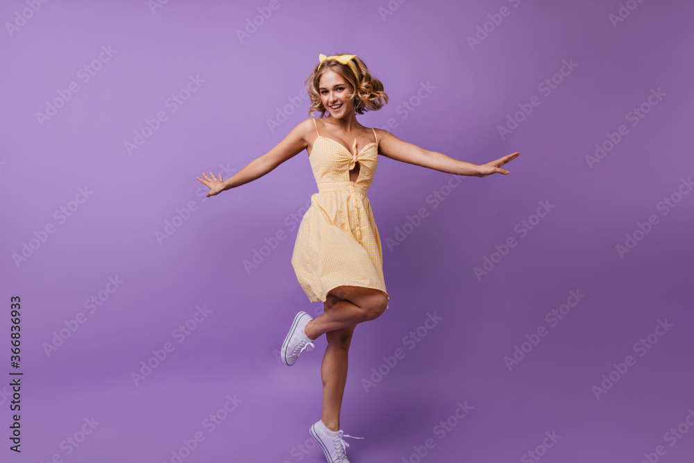 Full-length portrait of beautiful fair-haired female model dancing with pleasure. Indoor shot of jocund woman in yellow dress enjoying photoshoot.