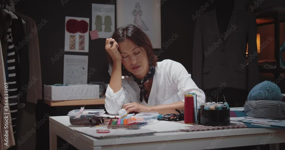 Tired fashion designer with brown hair fell asleep at tailor studio while working overtime. Mature woman sitting at table with dark atmosphere around. Tired fashion designer with brown hair fell asleep at tailor studio while working overtime. Mature woman sitting at table with dark atmosphere around.