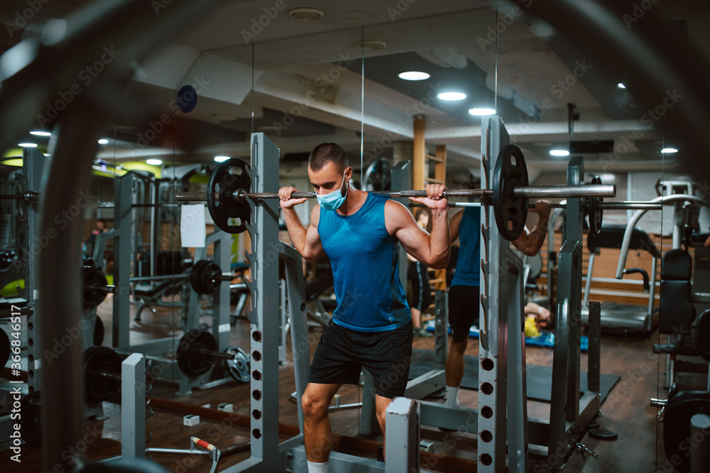 A young caucasian athlete man with a mask on his face exercises and lifts weights in the gym. COVID 19 coronavirus protection