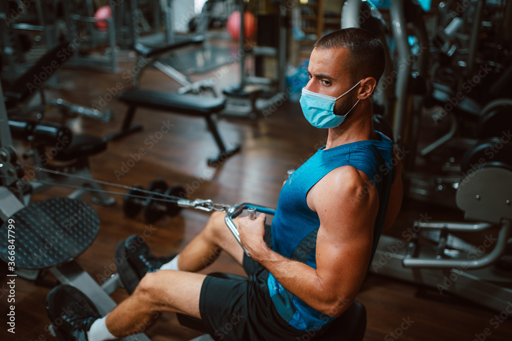 A handsome young caucasian man with a face mask is exercising in the gym on a device. Protection against COVID - 19 coronavirus A handsome young caucasian man with a face mask is exercising in the gym on a device. Protection against COVID - 19 coronavirus