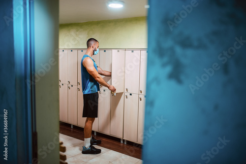 A young muscular caucasian man with a medical face mask takes clothes from the closet in the gym locker room. COVID 19 coronavirus protection