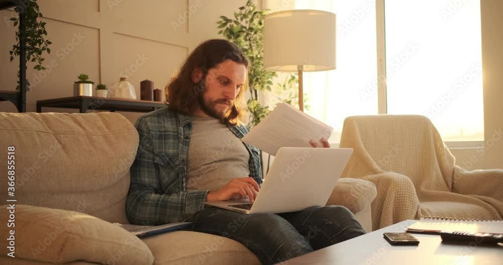 Man analyzing document and working on laptop sitting on sofa at home Man analyzing document and working on laptop sitting on sofa at home