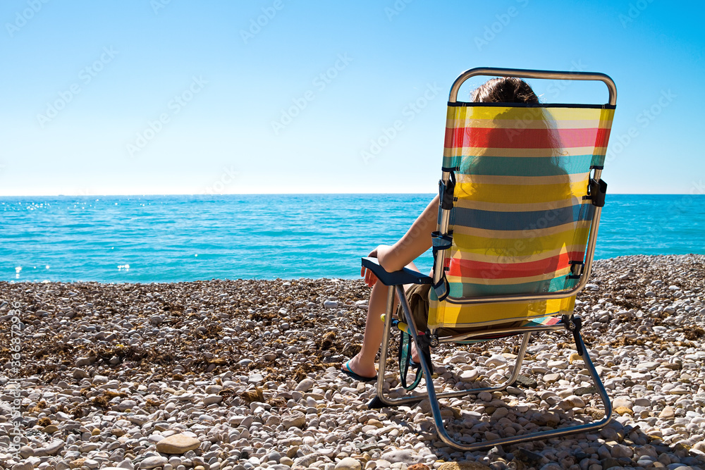 Long haired woman sunbathing in multi colored deck chair outdoor on pebble beach seaside view. Long haired woman sunbathing in multi colored deck chair outdoor on pebble beach seaside view.