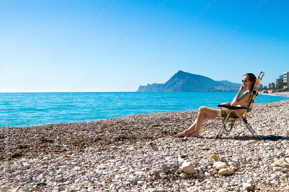 The beautiful woman sunbathing and sitting on a folding deck chair on the beach.