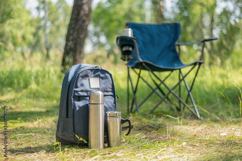 Tourist backpack, vacuum flask, chair and steel cup on green forest background.