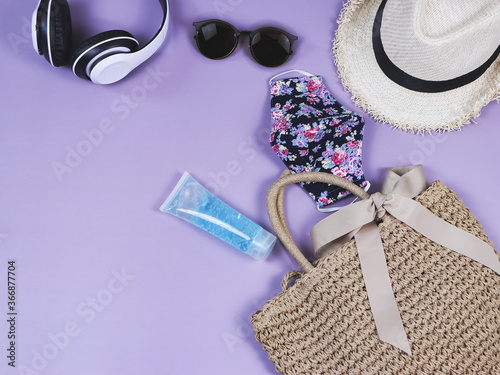 COVID-19 prevention , summer and new normal concept, top view of women's vacation accessories with flowery pattern fabric mask and sanitizer gel on purple background