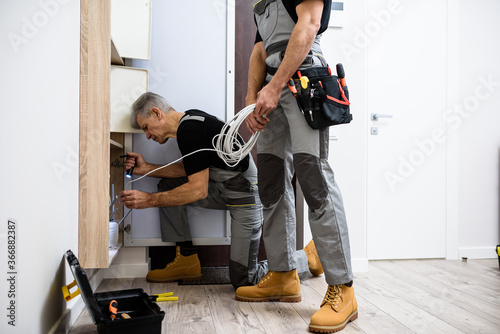 Aged electrician, repairman in uniform working, examining ethernet cable or router in fuse box using flashlight while his colleague holding cable
