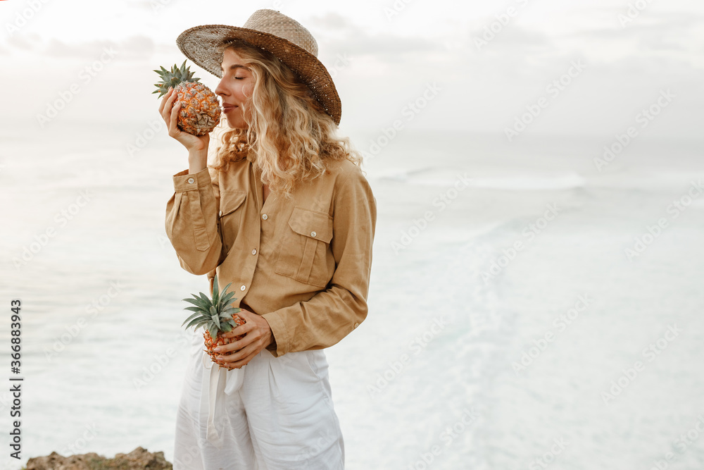 Beautiful woman with curly hair is holding pineapple fruit on the beach, smell tropical fruit