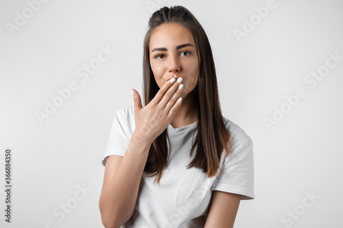 Pretty carefree modern young woman with long chestnut hair dressed in casual clothes covering mouth with one hand on white background. Keep silence concept