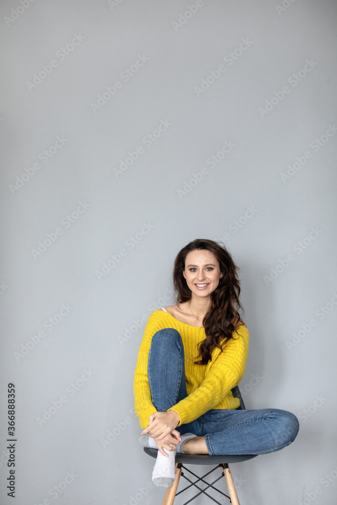 Beautiful young woman in casual clothes sits with her legs on a modern chair against a gray wall. Beautiful young woman in casual clothes sits with her legs on a modern chair against a gray wall.