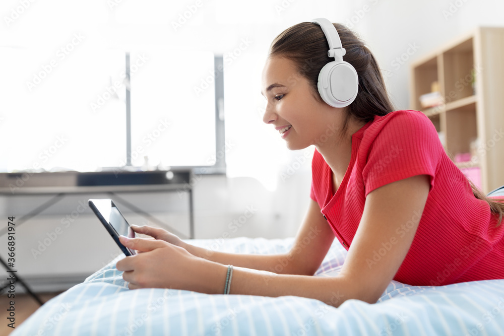 Naklejka premium children, technology and communication concept - smiling teenage girl in headphones listening to music on tablet computer at home