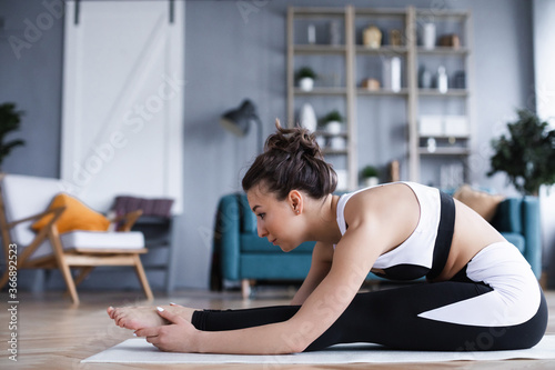 Beautiful woman with a good figure in sportswear doing stretching exercises while sitting on a yoga mat in the living room.