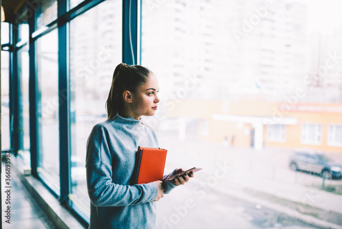 Bild auf Leinwand Beautiful hipster girl standing near window in modern loft office interior looking outside on rainy weather waiting for feedback from taxi service