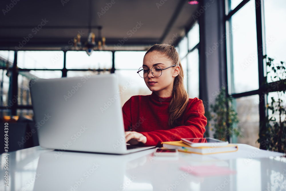 Gorgeous pensive professional female freelancer searching useful information on websites while working remotely on laptop computer.Young talented student dressed in red sweater using technology