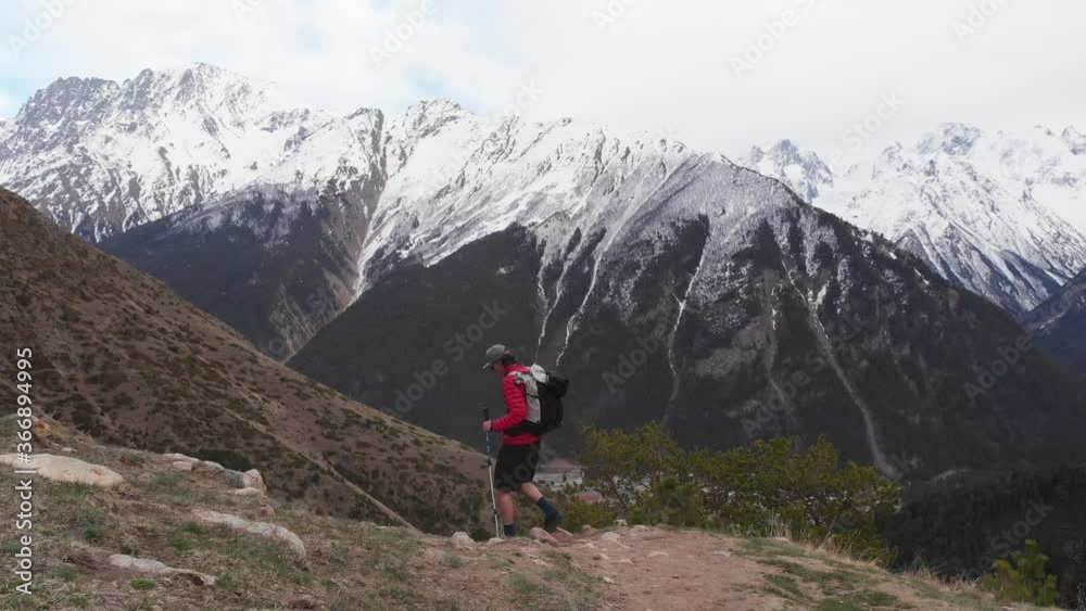 a young man in shorts sunglasses and a cap with a backpack goes along the route in the mountains with tracking sticks. Trekking and hiking in the mountains in the evening