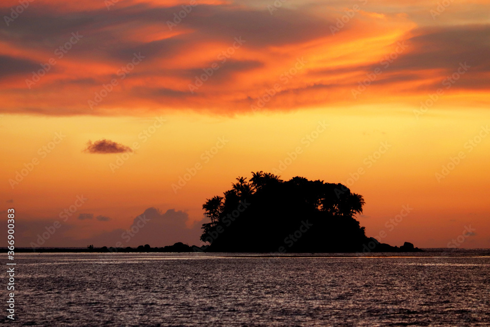 Sunset on a beach, silhouette of tropical island with palm trees in dark sea. Dramatic orange sky with clouds, romantic travel and vacation concept