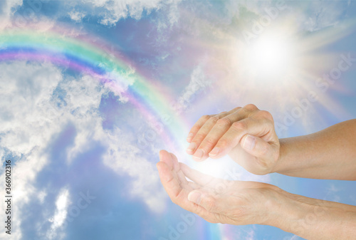 Capturing the light of a beautiful rainbow - female cupped hands with a ball of white light appearing to capture rainbow energy against a blue sky with fluffy clouds, bright sunburst and copy space 