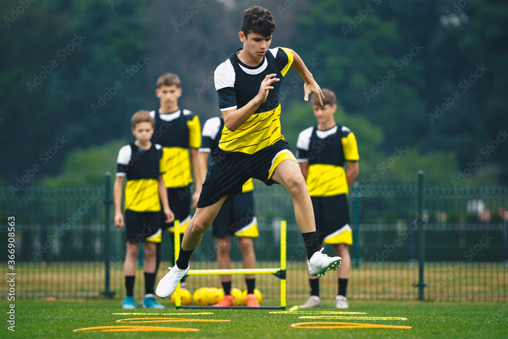 Naklejka premium Young Footballer on Training Jumping Over Obstacles. Summer Soccer Camp. Players in a Team Practicing and Improving Speed, Strength and Agility Skills