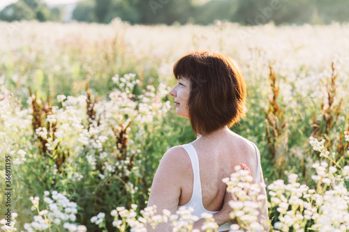 Middle aged mature red haired woman in white top with sensitive skin and freckles during yoga routine in sunny morning enjoys sunset in summer meadow field with chamomiles flowers.