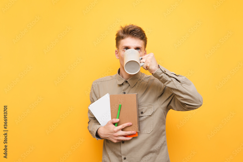 Funny student in a shirt and exercise books and books in his hands drinks a hot drink from a cup on a yellow background. Nerd student drinking tea from cup. Isolated. Copy space Funny student in a shirt and exercise books and books in his hands drinks a hot drink from a cup on a yellow background. Nerd student drinking tea from cup. Isolated. Copy space