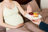 Close up of pregnant woman lying in bed don't want to eat a cake and makes stop gesture. No sweet and dessert during pregnancy concept