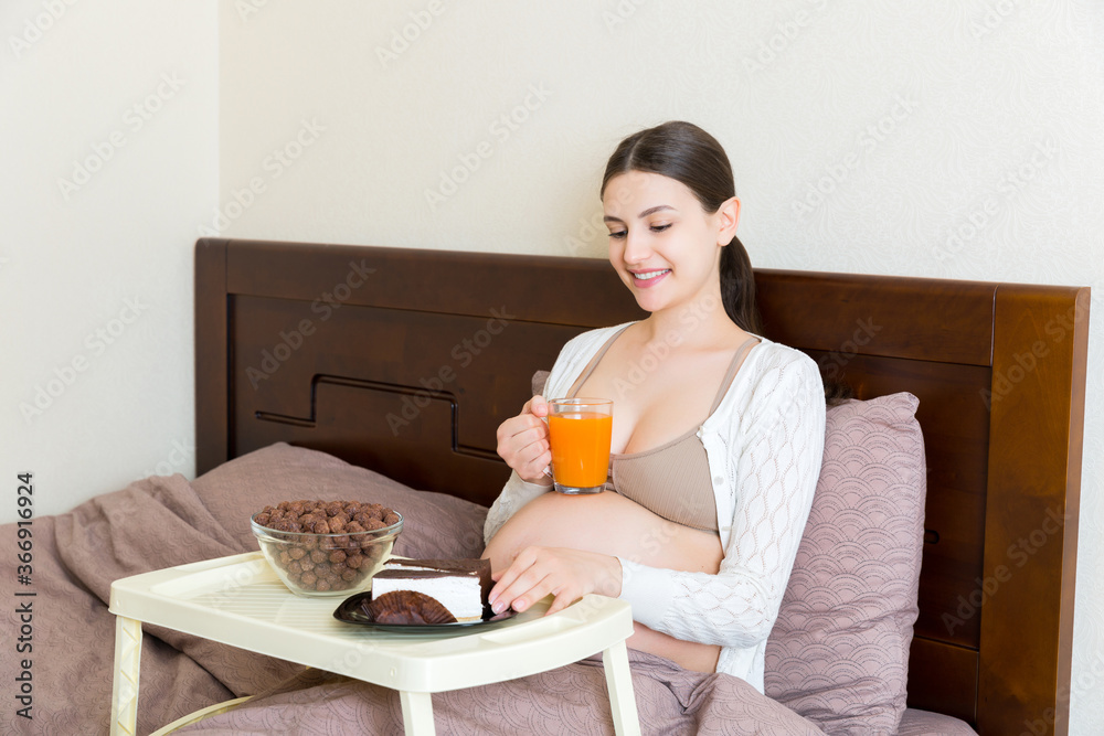 Pregnant woman is eating chocolate cereal balls, a piece of cake on breakfast tray and drinking juice in bed. Diet during pregnancy concept