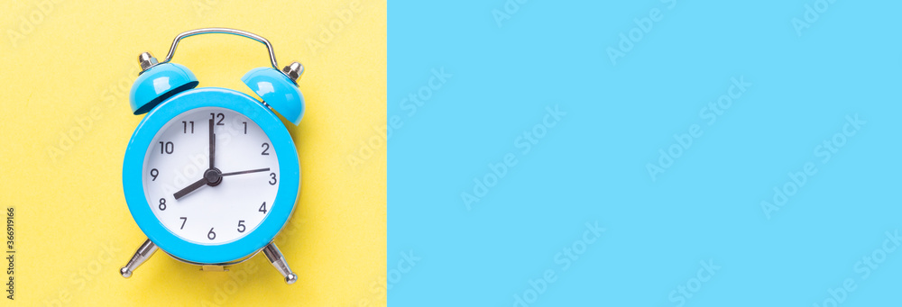 Blue alarm clock on two tone color yellow and blue background. Top view, place for text. Long banner Blue alarm clock on two tone color yellow and blue background. Top view, place for text. Long banner