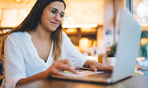 Charming caucasian female in trendy eyewear for vision correction sitting on cafe interior during remote job, positive woman blogger enjoying share multimedia content via modern laptop computer