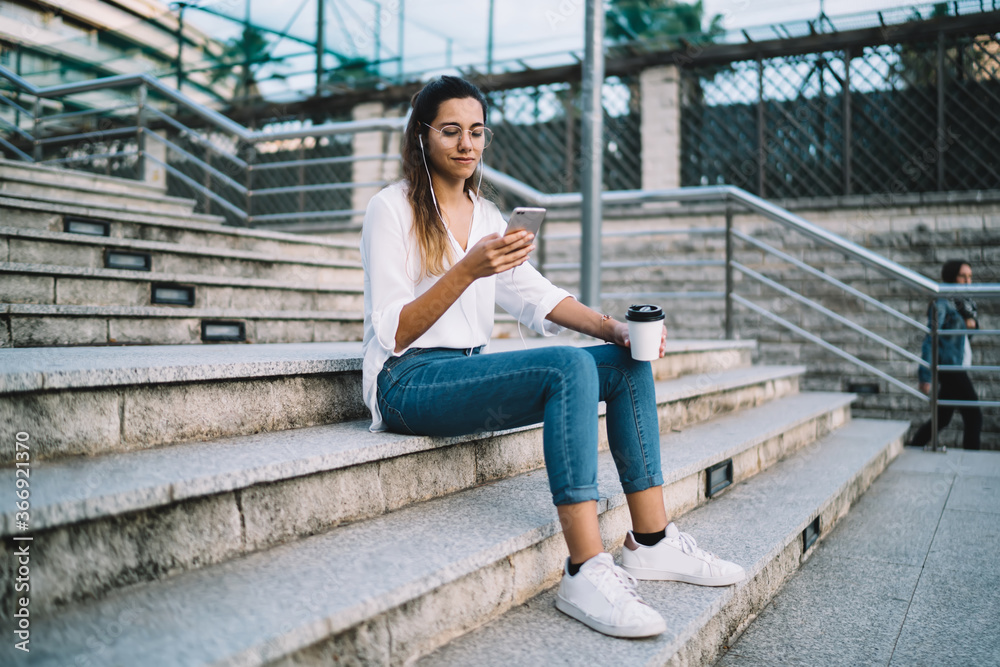 Positive caucasian woman meloman downloading music to mobile phone playlist sitting on stairs with coffee to go,cheerful female blogger listening music on smartphone share content in social networks