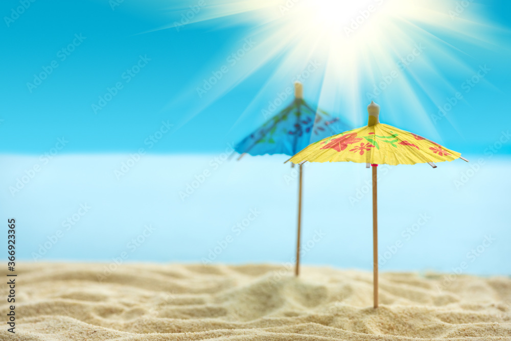 Fototapeta premium Sun umbrellas on sandy beach with blurry blue ocean, sky and sun beams. Summer background. Soft focus