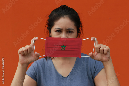 A woman with Morocco flag on hygienic mask in her hand and lifted up the front face on orange color background. Tiny Particle or virus corona or Covid 19 protection.