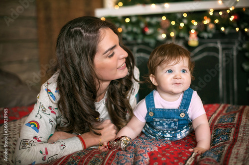 A beautiful young mother lies on the bed with her little daughter against the background of the Christmas fireplace. The woman looks at the child and smiles.