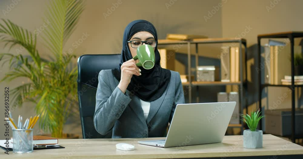 Young Arabic woman in hijab and glasses sitting at desk in office and ...