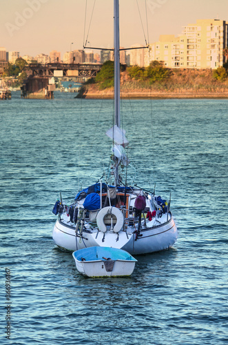 Photography The back of a sail boat / yacht moored in the harbour - wet clothes hanging off the side - sails down - dinghy tided to it - in the shade at sunset
