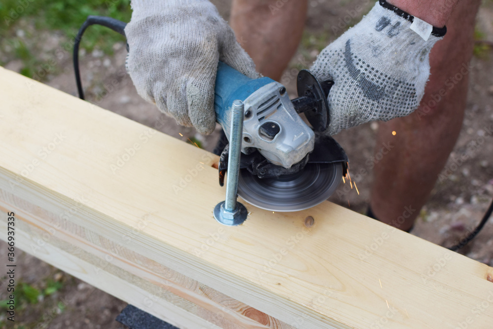 Craftsman hand sawing metal screw with disk grinder on wooden plank outdoors with sparks around.
