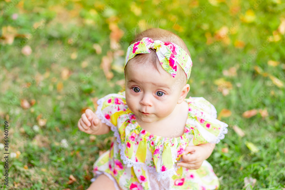 portrait of a small baby girl 7 months old sitting on the green grass in a yellow dress, walking in the fresh air