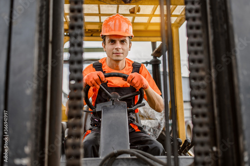 Front view of a serious face young caucasian man builder in overalls and orange protective helmet drive a construction loader