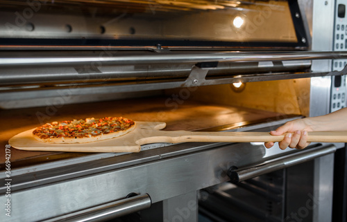 Close up baker is holding a wood peel with fresh pizza in an oven at a baking manufacture factory.
