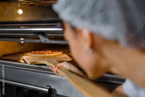 Young caucasian woman baker is holding a wood peel with fresh pizza and put it in an oven at a baking manufacture factory.
