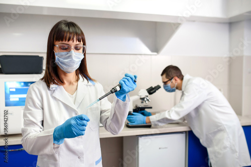 In the hospital laboratories working process  a young woman doctor is using micropipette test tubes  in the background a laboratory assistant is looking at the medical microscope