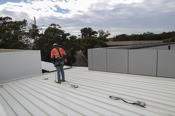 Trained competent industrial construction worker wearing safety helmet fall arrest harness and clipping Carabiner on roof anchor point while working near open edge as fall restraint protection     