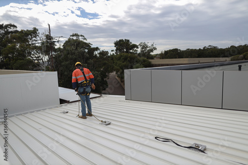 Trained competent industrial construction worker wearing safety helmet fall arrest harness and clipping Carabiner on roof anchor point while working near open edge as fall restraint protection     